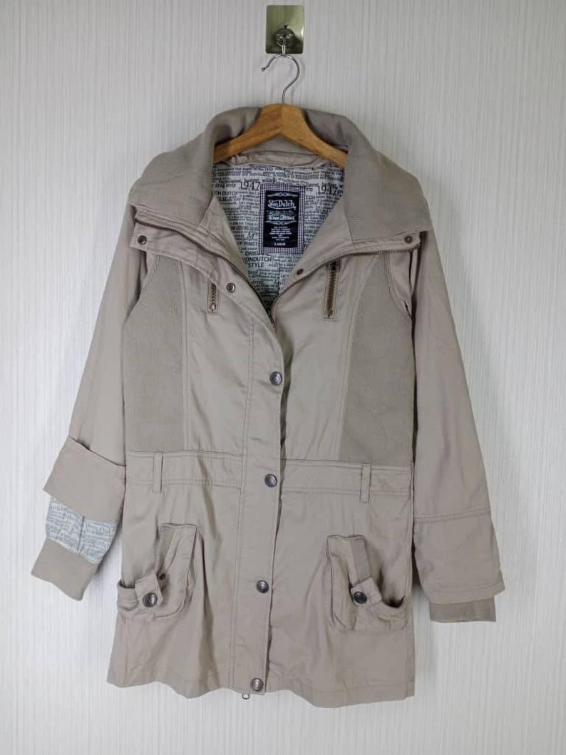 Peut inclure: Une veste beige &agrave; manches longues avec un col montant, une fermeture &eacute;clair et plusieurs poches. La veste est doubl&eacute;e d'une doublure imprim&eacute;e avec le texte "1947" et "Love & Luck".