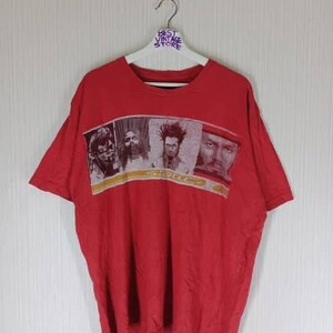 Puede incluir: Camiseta roja con un gráfico que presenta cuatro caras y la palabra "Static" en una banda horizontal en el pecho. La camiseta está colgada en una percha blanca contra una pared blanca.