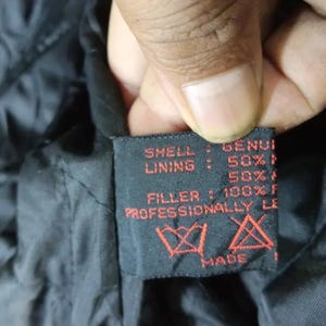 May include: Close-up of a black garment tag with red text. The tag reads "SHELL: GENUI, LINING: 50%, FILLER: 100% F, PROFESSIONALLY LE, MADE". The tag also features care symbols.