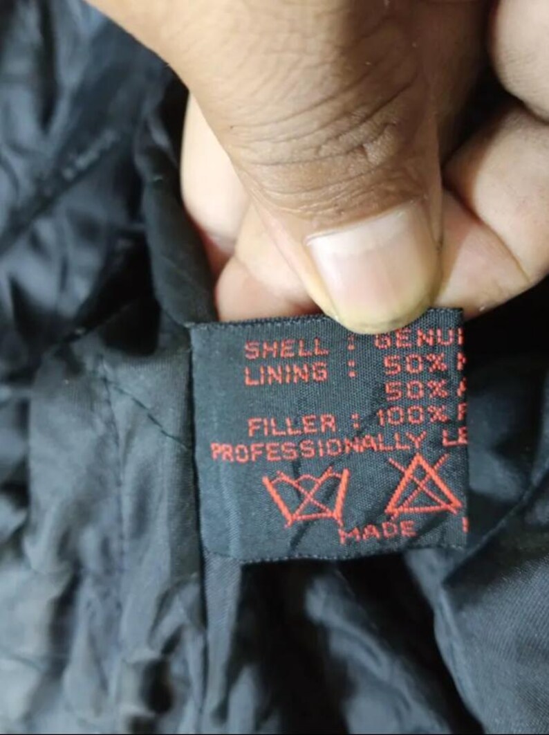 May include: Close-up of a black garment tag with red text. The tag reads "SHELL: GENUI, LINING: 50%, FILLER: 100% F, PROFESSIONALLY LE, MADE". The tag also features care symbols.