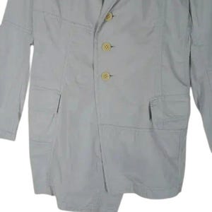 May include: A light gray blazer with three beige buttons and two front pockets. The blazer has a notched lapel and long sleeves. The seams are visible, adding a unique design element to the garment.