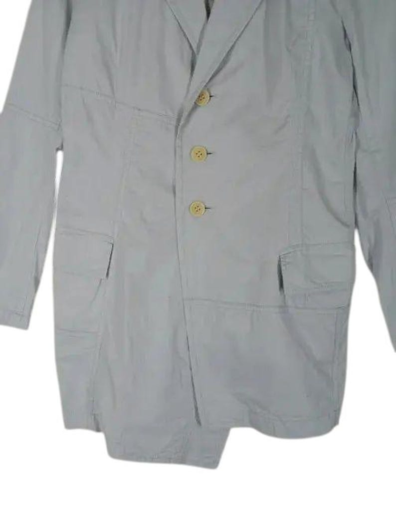 May include: A light gray blazer with three beige buttons and two front pockets. The blazer has a notched lapel and long sleeves. The seams are visible, adding a unique design element to the garment.