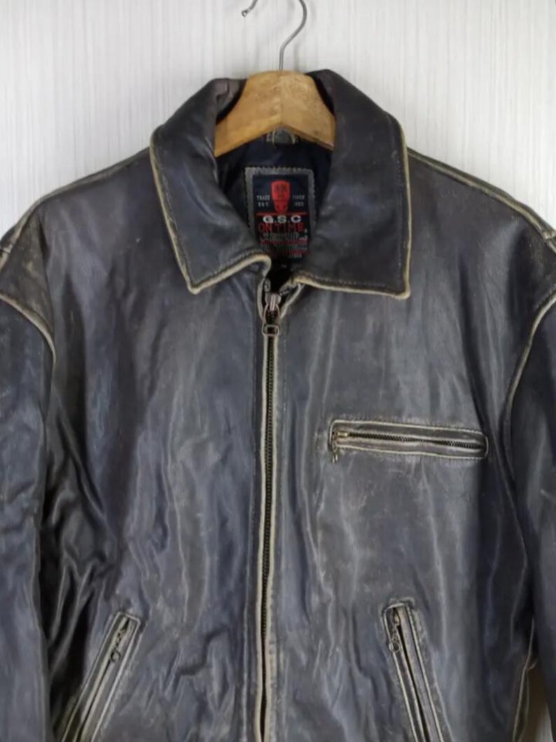 May include: A distressed, dark gray leather jacket with a zip-up front and multiple pockets. The jacket features a classic collar and a weathered appearance, suggesting vintage styling. The G.S.C. brand label is visible on the inside.