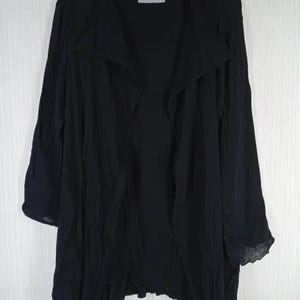 Op de afbeelding: Zwarte jas met open voorkant en lange mouwen. De jas heeft een gedrapeerde kraag en een asymmetrische zoom. De stof lijkt licht en licht getextureerd. De jas hangt aan een houten kleerhanger.