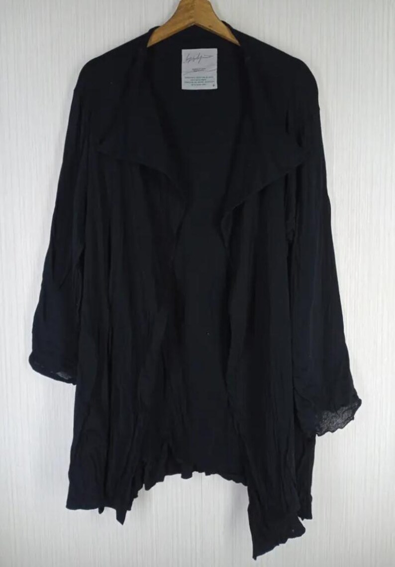 Op de afbeelding: Zwarte jas met open voorkant en lange mouwen. De jas heeft een gedrapeerde kraag en een asymmetrische zoom. De stof lijkt licht en licht getextureerd. De jas hangt aan een houten kleerhanger.