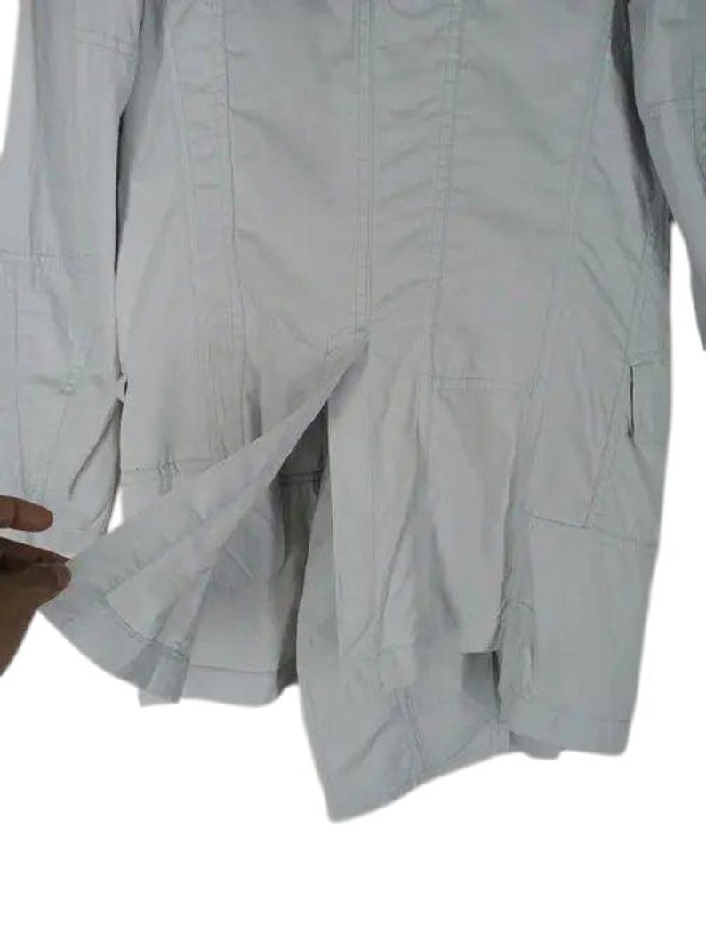 May include: A light gray jacket with a tailored design. The jacket features a button closure, multiple pockets, and a split hem. The fabric appears to be lightweight, suitable for transitional weather.