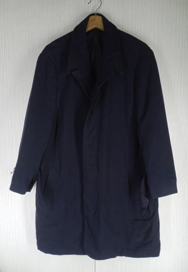 Pu&ograve; includere: Un cappotto blu navy con colletto a revers e due tasche anteriori. Il cappotto &egrave; appeso a una gruccia di legno su uno sfondo bianco. Il cappotto sembra essere fatto di un tessuto pesante.