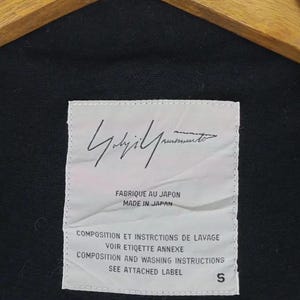 Op de afbeelding: Zwart kledingstuk met een wit label met de handtekening van de ontwerper en tekst. Het label luidt "FABRIQUE AU JAPON MADE IN JAPAN" en bevat wasvoorschriften. De maat wordt aangegeven als "S".