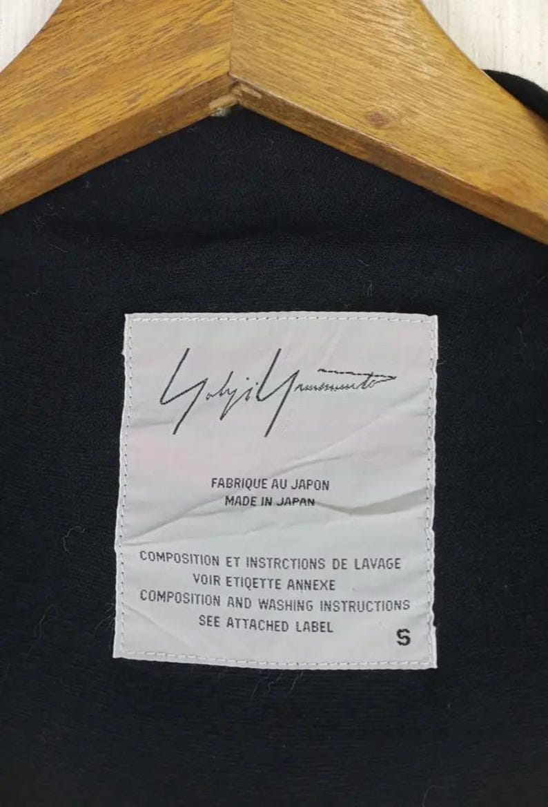 Op de afbeelding: Zwart kledingstuk met een wit label met de handtekening van de ontwerper en tekst. Het label luidt "FABRIQUE AU JAPON MADE IN JAPAN" en bevat wasvoorschriften. De maat wordt aangegeven als "S".