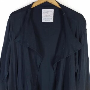 Op de afbeelding: Een zwarte jas met open voorkant en brede reverskraag, hangend aan een houten hanger. De jas heeft lange mouwen en een label met witte tekst. De stof lijkt licht van gewicht. De algehele stijl is minimalistisch en modern.