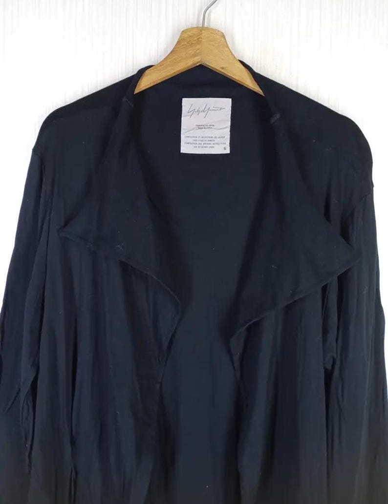 Op de afbeelding: Een zwarte jas met open voorkant en brede reverskraag, hangend aan een houten hanger. De jas heeft lange mouwen en een label met witte tekst. De stof lijkt licht van gewicht. De algehele stijl is minimalistisch en modern.