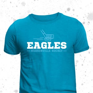 May include: A turquoise blue t-shirt with a white hockey stick and puck graphic. The text "EAGLES - HUDSONVILLE HOCKEY -" is printed in white on the shirt.