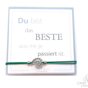 Könnte beinhalten: Ein grünes Gummiband mit einem silbernen Anhänger in Form einer Muschel. Das Armband hat eine Karte daran befestigt, auf der steht "Du bist das BESTE was mir je passiert ist."