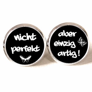 Könnte beinhalten: Ein Paar schwarze Knopf-Ohrringe mit silbernen Einstellungen. Der linke Ohrring sagt "nicht perfekt" mit einer Schmetterlingsgrafik. Der rechte Ohrring sagt "aber einzig artig!" mit einer Schmetterlingsgrafik.