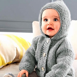 Può includere: Un cardigan grigio a maglia con cappuccio e chiusura a bottoni, indossato da un bambino seduto su una superficie bianca.