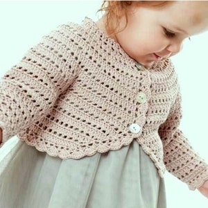 Op de afbeelding: Een lichtroze gehaakte trui met een gegolfde zoom en knoopsluiting. De trui wordt gedragen door een klein kind.