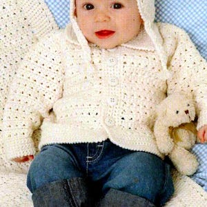 May include: A baby wearing a cream-colored, crocheted hat and cardigan with button closures, paired with blue denim jeans. The baby is holding a small stuffed animal. The outfit is on a white crocheted blanket.