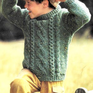 Op de afbeelding: Een groene kabelgebreide trui met een ronde hals. De trui wordt gedragen door een kind dat op een houten bank staat. Het kind draagt een bruine hoed en een bruine broek.