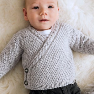 May include: A baby wearing a gray knitted cardigan with a wrap-around style and button closures. The cardigan has a textured knit pattern.
