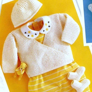 Op de afbeelding: Een witte gebreide baby-outfit met een bijpassende muts, een geel gestreepte romper en een kleine teddybeer. De outfit heeft een witte kraag met een bloemenpatroon.