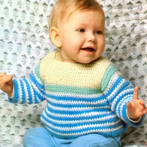 Puede incluir: Un bebé con un suéter de crochet a rayas con rayas amarillas, verdes y azules. El suéter tiene un cuello de crochet y está hecho con hilo blanco.
