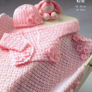 May include: A pink crocheted baby blanket, hat, and cardigan set with a crocheted flower design. The set is laid out on a white surface with a pink teddy bear. The text "double knit" and "36 - 56 cm, 14 - 22 in" is visible in the image. Crochet.