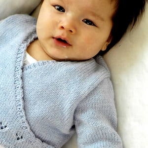 Può includere: Un cardigan di maglia blu chiaro per neonato con uno stile avvolgente e maniche lunghe. Il cardigan ha un delicato motivo a pizzo sui bordi.