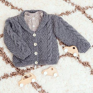 May include: A gray cable-knit cardigan with a collared shirt underneath, and three small wooden toy cars with white wheels. The cardigan has a button-up front and a textured knit design. The shirt has a brown and white plaid pattern.