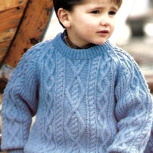 May include: A young boy wearing a light blue cable knit sweater. The sweater has a classic cable knit pattern and a crew neck.