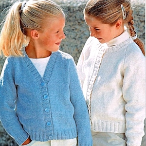 Könnte beinhalten: Zwei junge Mädchen tragen Strickjacken. Eine ist hellblau mit passenden Knöpfen, die andere ist weiß. Beide Strickjacken haben einen V-Ausschnitt. Die Mädchen schauen sich an.