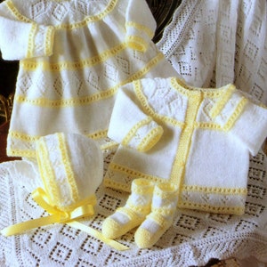 May include: A white knitted baby outfit with yellow trim, including a dress, cardigan, hat, and booties. The outfit is displayed on a white crocheted blanket.
