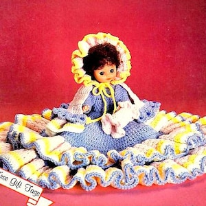 May include: A handmade doll with a blue crocheted dress and a yellow-trimmed bonnet. The doll is holding a small white crocheted item. The doll is surrounded by a large, ruffled skirt in yellow, white, and blue. The image includes the text "Free Gift Tags".