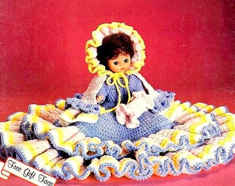 May include: A handmade doll with a blue crocheted dress and a yellow-trimmed bonnet. The doll is holding a small white crocheted item. The doll is surrounded by a large, ruffled skirt in yellow, white, and blue. The image includes the text "Free Gift Tags".