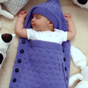 May include: A purple knitted baby sleeping bag with a hood and buttons down the side. The baby is sleeping soundly inside the bag.