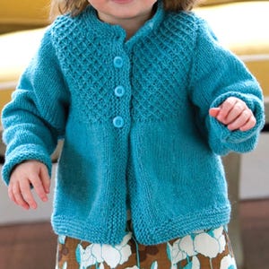 Puede incluir: Un cárdigan de punto turquesa con canesú texturizado y tres botones a juego. Las mangas son largas y el cárdigan parece hecho a mano. La prenda está diseñada para un niño.