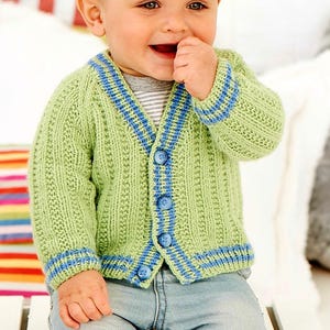 Può includere: Un cardigan da bambino lavorato a maglia verde chiaro con strisce blu sui polsini, sul colletto e sull'abbottonatura. Il cardigan ha uno scollo a V ed è chiuso con quattro bottoni blu. Il bambino indossa jeans blu e una maglietta a righe grigie.