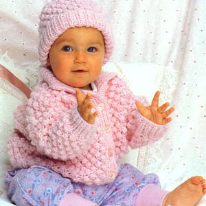 May include: A pink knitted baby cardigan with a matching hat. The cardigan has a button fastening and a textured stitch pattern. The baby is wearing the cardigan and hat and is sitting on a white surface.