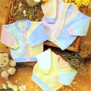 May include: Several pastel-colored knitted baby sweaters and a matching top are arranged on a wicker basket and wooden surface. The sweaters feature blue, pink, and yellow panels with button closures. A teddy bear and flowers are also present.