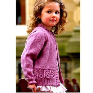 May include: A young girl wearing a pink knitted cardigan with a decorative lace trim. The cardigan has a button closure and long sleeves.