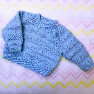 May include: A light blue knitted baby sweater with a button closure. The sweater has a textured pattern and is made of soft yarn.