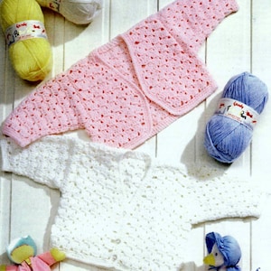 May include: Two crocheted baby cardigans, one pink and one white, laid out on a white surface. The cardigans are made with a simple stitch pattern and have a button closure. The image also includes balls of yarn in pink, white, and blue.