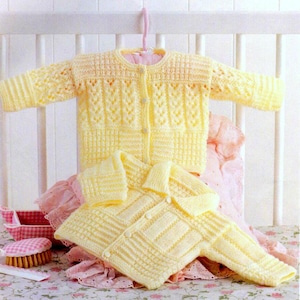 May include: Two yellow knitted baby cardigans. One is hanging on a pink hanger, the other is folded below. The cardigans have button closures and textured patterns. The background includes a pink dress and a crib.