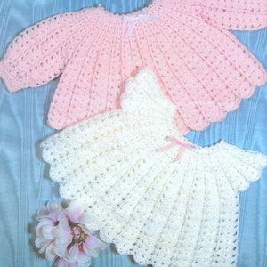 May include: Two crocheted baby garments, one a pink sweater and the other a white dress. Both garments are made with a delicate, lacy stitch.