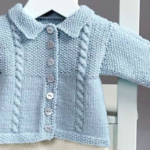 Może przedstawiać: Jas dla niemowlęcia w jasnoniebieskim kolorze, z białym kołnierzykiem i zapięciem na guziki. Sweter ma wzór warkoczy i jest pokazany na białym drewnianym wieszaku.