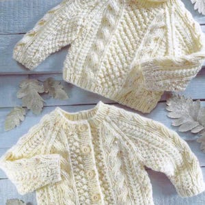 Op de afbeelding: Twee crèmekleurige gebreide babyvestjes met een kabelbreipatroon en knopen.