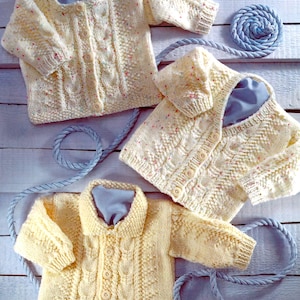 Könnte beinhalten: Drei gelbe gestrickte Baby-Cardigans mit Knöpfen. Die Cardigans haben ein Zopfmuster und liegen auf einer weißen Holzoberfläche.