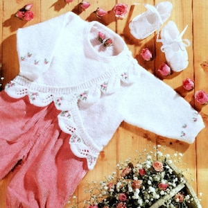 May include: White knitted baby cardigan with a scalloped edge and embroidered flowers. The cardigan is laid out on a wooden surface with pink flowers and a basket of white flowers.