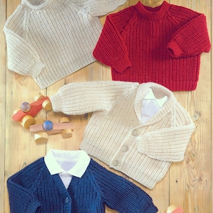 Op de afbeelding: Vier gebreide kindertruien in crème, rood en marineblauw. De truien omvatten een ronde hals, een vest met knopen en een vest met kraag. Houten speelgoedauto's zijn ook zichtbaar op het houten oppervlak.