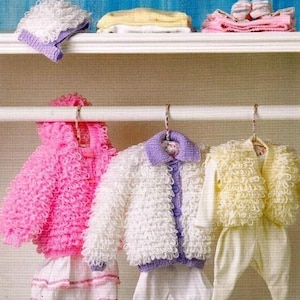 May include: Three knitted baby sweaters hanging on a white shelf. The sweaters are pink, white, and yellow. The sweaters are all made with a fluffy, textured yarn.