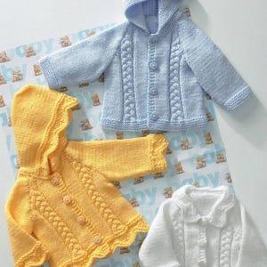 Może przedstawiać: Trzy dzianinowe sweterki dla niemowląt w kolorze niebieskim, żółtym i białym. Niebieskie i żółte sweterki mają kaptury i zapięcia na guziki. Biały sweterek ma kołnierzyk. Wszystkie trzy sweterki mają wzór warkocza. Sweterki są prezentowane na wzorzystej powierzchni.